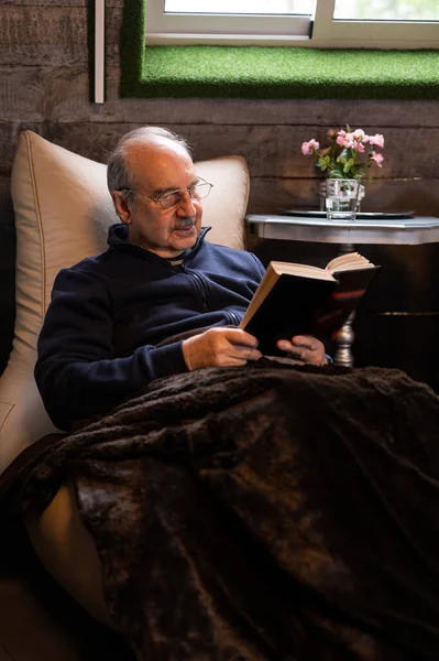 Elderly man with a mustache and reading glasses covered with a soft warm blanket sit on a bean bag and read a book in cozy interior of wooden house