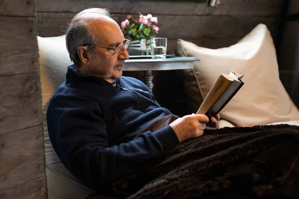 Elderly man with a mustache and reading glasses covered with a soft warm blanket sit on a bean bag and read a book in cozy interior of wooden house