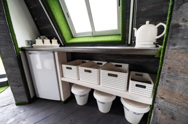 Small kitchen elements in wooden summer house. Refrigerator, kettle, hot drinks, crockery and recycling cans in small kitchen. Original, unusual, country interior architecture designs example.