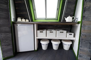 Small kitchen elements in wooden summer house. Refrigerator, kettle, hot drinks, crockery and recycling cans in small kitchen. Original, unusual, country interior architecture designs example.