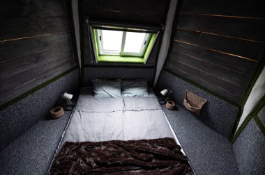 Double bed on carpet floor in bedroom of wooden cabin. Pyramid shape bungalow from inside. Small room, angled walls, window light, made bed. Handmade interior of wooden house.