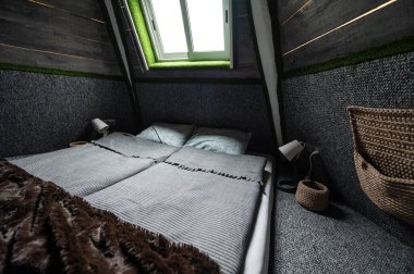 Double bed on carpet floor in bedroom of wooden cabin. Pyramid shape bungalow from inside. Small room, angled walls, window light, made bed. Handmade interior of wooden house.