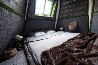 Double bed on carpet floor in bedroom of wooden cabin. Pyramid shape bungalow from inside. Small room, angled walls, window light, made bed. Handmade interior of wooden house.