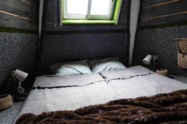 Double bed on carpet floor in bedroom of wooden cabin. Pyramid shape bungalow from inside. Small room, angled walls, window light, made bed. Handmade interior of wooden house.