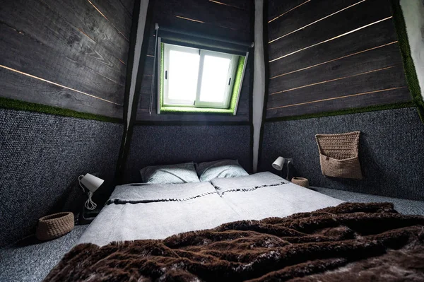Double bed on carpet floor in bedroom of wooden cabin. Pyramid shape bungalow from inside. Small room, angled walls, window light, made bed. Handmade interior of wooden house.