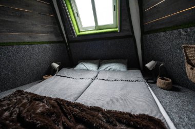 Double bed on carpet floor in bedroom of wooden cabin. Pyramid shape bungalow from inside. Small room, angled walls, window light, made bed. Handmade interior of wooden house.
