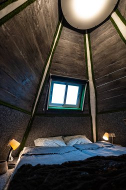 Double bed in bedroom of pyramid shape wooden cabin. Pyramid shape walls of bungalow from inside. Small room, angled walls, window light, ceiling lamp, tree trunks in the corners, made bed.