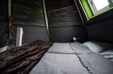 Double bed on carpet floor in bedroom of wooden cabin. Pyramid shape bungalow from inside. Small room, angled walls, window light, made bed. Handmade interior of wooden house.
