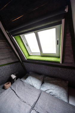 Double bed on carpet floor in bedroom of wooden cabin. Pyramid shape bungalow from inside. Small room, angled walls, window light, made bed. Handmade interior of wooden house.