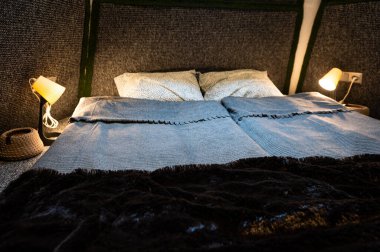 Double bed on carpet floor in bedroom of wooden cabin. Pyramid shape bungalow. Small room, angled walls, window light, made bed, night lamp and knitted basket. Handmade interior of wooden house.