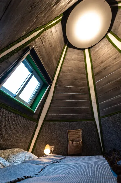 Double bed in bedroom of pyramid shape wooden cabin. Pyramid shape walls of bungalow from inside. Small room, angled walls, window light, ceiling lamp, tree trunks in the corners, made bed.