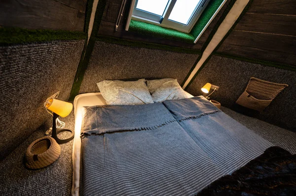 Double bed on carpet floor in bedroom of wooden cabin. Pyramid shape bungalow from inside. Small room, angled walls, window light, made bed. Handmade interior of wooden house.