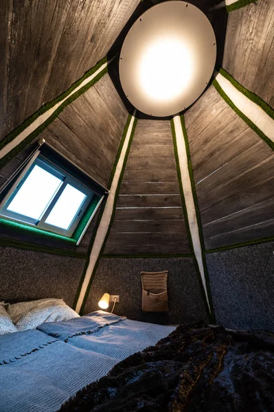 Double bed in bedroom of pyramid shape wooden cabin. Pyramid shape walls of bungalow from inside. Small room, angled walls, window light, ceiling lamp, tree trunks in the corners, made bed.