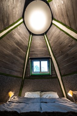 Double bed in bedroom of pyramid shape wooden cabin. Pyramid shape walls of bungalow from inside. Small room, angled walls, window light, ceiling lamp, tree trunks in the corners, made bed.