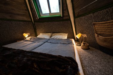 Double bed in bedroom of pyramid shape wooden cabin. Pyramid shape walls of bungalow from inside. Small room, angled walls, window light, ceiling lamp, tree trunks in the corners, made bed.