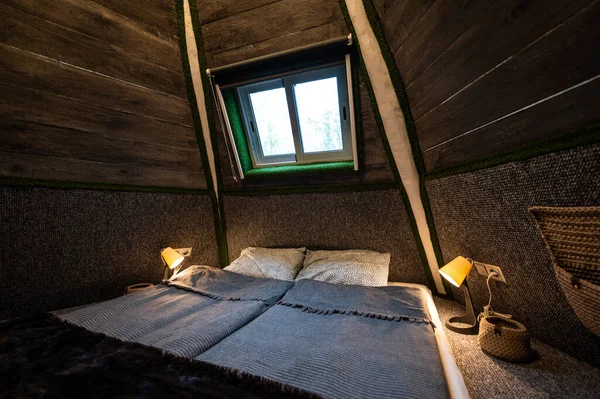 Double bed in bedroom of pyramid shape wooden cabin. Pyramid shape walls of bungalow from inside. Small room, angled walls, window light, ceiling lamp, tree trunks in the corners, made bed.