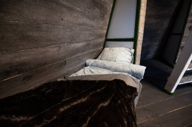 Single bed with clean linen in bedroom niche of wooden cabin. Pyramid shape bungalow summer house. Angled surrounded walls, gray horizontal wooden wall planks. Nonstandard bed frame construction.