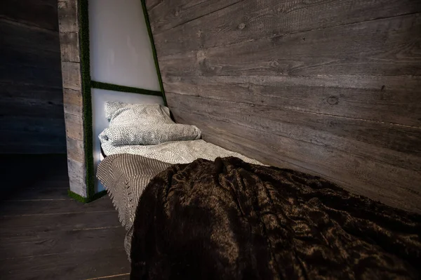 Single bed with clean linen in bedroom niche of wooden cabin. Pyramid shape bungalow summer house. Angled surrounded walls, gray horizontal wooden wall planks. Nonstandard bed frame construction.