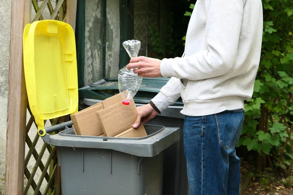 A man sorting waste. A person throwing cardboard packaging and a plastic bottle into a yellow recycling bin.