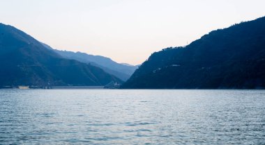 Layers of blue mountains rise above the lake, bathed in blue light and sunlight. The calm water reflects the beauty of the surroundings in Tehri Lake. clipart