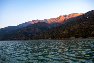 Sahne, dağın tepelerini aydınlatan altın güneş ışığıyla gölü yakalıyor. Sakin sular Tahri Gölü 'nü çevreleyen yeşil dağların güzelliğini yansıtıyor.