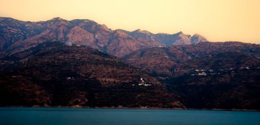 The sunset casts a warm glow over the mountains and small towns on the slopes. Smoke rises from the mountains, adding to the picturesque view of white houses and other structures at Tehri Lake. clipart