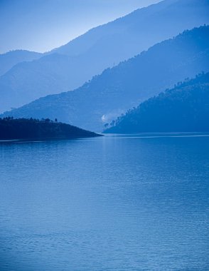 Blue mountains and mist create a serene backdrop at Tehri Lake during sunrise. The mountains are covered with trees and plants, adding to the natural beauty of the landscape, while the calm waters reflect the tranquility of the scene. clipart