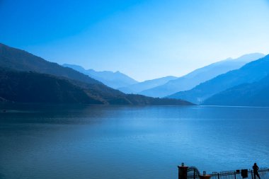 Bir adam mavi gölü ve katmanlı dağları gözlemliyor, diğer tarafta güzel bir gündoğumu var. Bu sakin manzara bölgenin doğal güzelliğini gözler önüne seriyor..