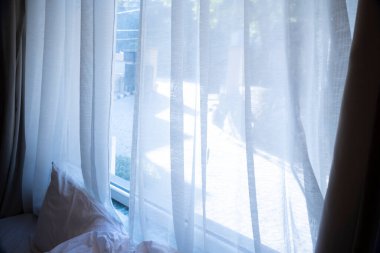 Beyaz perdeler oturma odasının penceresini süslüyor ve doğal ışığın içeri sızmasını sağlıyor. Pencerenin üzerinde bir yastık duruyor, dekora samimi bir dokunuş katıyor..