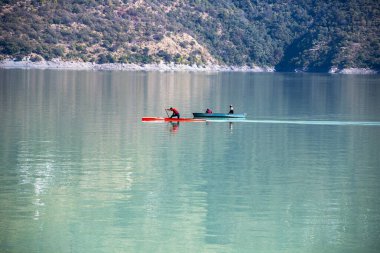 In calm waters, two men wait in one boat while another man rows in a kayaking boat. The peaceful setting at Tehri Lake enhances the enjoyment of their outing. clipart