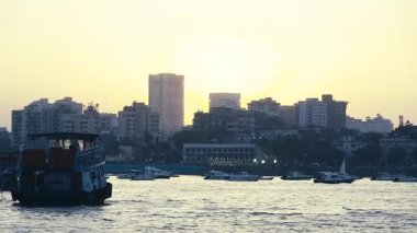 Mumbai şehrinin ufuk çizgisi deniz boyunca uzanır, büyüleyici bir manzara sunar. Birçok tekne ve feribot kıyıya yaklaşıyor, bu da aktif deniz hareketini gösteriyor. Şehrin arkasında parlayan güneş manzaranın güzelliğini arttırır..