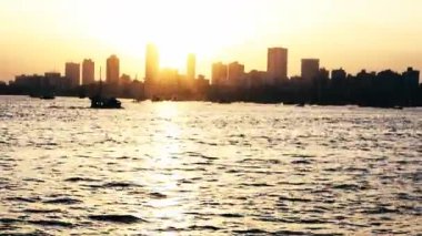 Mumbai şehrinin ufuk çizgisi deniz boyunca çarpıcı bir manzara sunuyor. Pahalı bir atmosfer yaratıyor. Denizde gezinen tekneler, sakin kıyı manzarasını güzelleştirir. Güneş şehrin arkasında ışıl ışıl parlıyor, manzaraya sıcaklık katıyor..