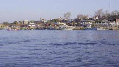Mathura 'nın Vrindavan kentindeki Yamuna Nehri bankasında kenetlenmiş tekneler ve park halindeki araçlar yer alıyor. Bu sahne bölgenin popülerliğini ve ulaşılabilirliğini vurguluyor.
