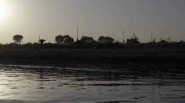 Nehir kıyılarının karanlık silueti uzun otlarla çevrili birkaç kişi tarafından karşıt görülür. Bu sahne su kenarında sakin bir anı yakalar..