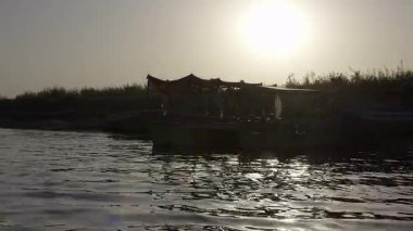 Birkaç kişi, uzun otlarla çevrili nehir kıyılarının karanlık siluetine karşı durmaktadır. Sudaki tekneler, genel sahneye derinlik katıyor..