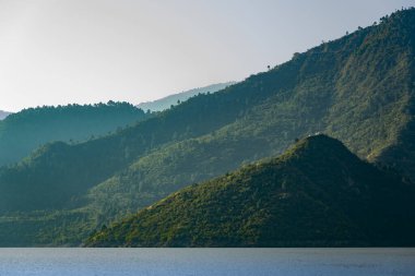 Yeşil kaplı dağlar, aşağıdaki bir gölün üzerinde görkemli bir şekilde yükseliyor. Sabah ışıkları dağları aydınlatır, doğanın güzelliğinin çarpıcı bir görsel görüntüsünü yaratır..