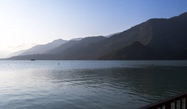 Mavi sis, sabah ışığında dağları kaplıyor. Aşağıda mavi bir göl var. Işık dalgaları yansıtır ve bulutlu bir gökyüzünün altındaki evlerden duman yükselir..