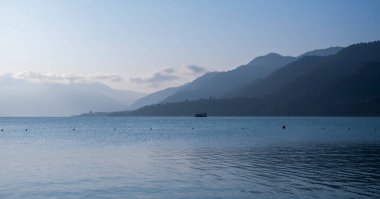 Mavi sis, sabah ışığında dağları kaplıyor. Aşağıda mavi bir göl var. Işık dalgaları yansıtır ve bulutlu bir gökyüzünün altında yüzen bir otel görünür..