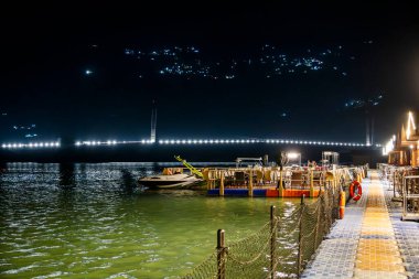 Tihri, Hindistan, 08 Aralık 2025: Kenetlenmiş tekneler göl suyunda dinlenirken, bir tarafta aydınlık bir tatil köyü görülebilir. Diğer taraftaki aydınlatılmış bir köprü tüm atmosferi güçlendirir..