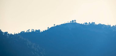 Çarpıcı bir günbatımı mavi dağları aydınlatırken, ağaçlar ve elektrik direkleri dağın zirvesinde dik duruyorlar. Sahne huzur ve güzellik saçıyor..