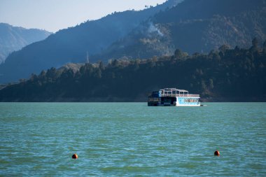 Tihri, Hindistan, 08 Aralık 2025: Göldeki bir mavnada yüzen bir otel, yakındaki evlerden yükselen dumanlar ve dağların çarpıcı manzarasını sunuyor. Sahne rahatlık ve doğayı harmanlıyor..