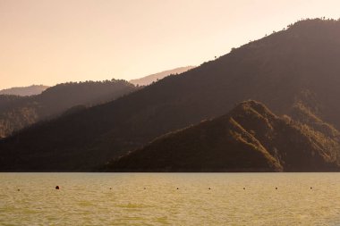 Güneşin kahverengi ışığı dağların üzerine ve aşağıdaki göle sıcak bir parıltı saçar. Bu büyüleyici sahne manzaranın doğal güzelliğini vurgular..