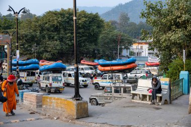 Rishikesh, Hindistan, 08 Aralık 2024: Yaşlı bir aziz, yakınlarda çay içen bir adam olarak yürür. Üstünde sallar olan cipler onları Rishikesh 'teki rafting istasyonuna götürmeye hazır..