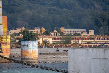 Rishikesh, Hindistan, 08 Aralık 2024: Rishikesh 'te Ganj Nehri' nin karşısında bir aşram bulunabilir, burada çaydanlıklar mevcuttur ve erkekler yürümektedir. Atmosfer huzuru yansıtıyor..