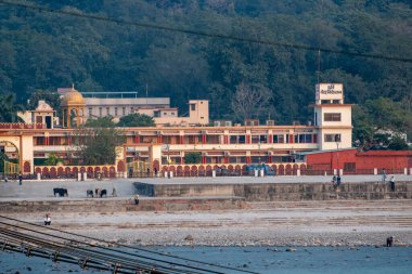 Rishikesh, Hindistan, 08 Aralık 2024: Rishikesh 'te Ganj Nehri' nin karşısındaki bir aşramda çaydanlıklar ve yürüyen adamlar ve yakınlarda oturan insanlar yer alıyor. Çevre huzurlu ve davetkar..