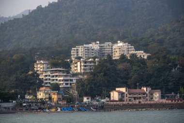 Rishikesh, Hindistan, 08 Aralık 2024: Ganj Nehri 'nin karşısındaki Rishikesh kasabası diğer taraftaki salları, insanları ve araçları gösteriyor. Bu sahne bölgenin canlı atmosferini yakalar..
