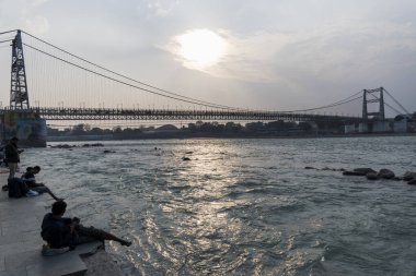 Rishikesh, Hindistan, 08 Aralık 2024: İnsanlar Ganj Nehri 'ne bakan Rishikesh' teki Janki Setu köprüsü yakınlarındaki ghat üzerinde oturuyorlar. Bu barışçıl ortam yansımayı ve rahatlamayı davet ediyor..