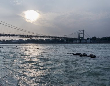 Akan nehir ve Rishikesh kasabası bulutlu bir gökyüzünün altında Janki Setu köprüsü tarafından çerçevelenmiştir. Bu sahne bir huzur ve güzellik hissi uyandırıyor..
