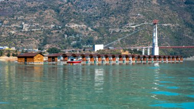 Gölde yüzen barakalar, arka planda bir köprü ile dağların çarpıcı bir görüntüsünü sağlar..