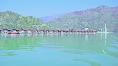 Tehri, Hindistan - 08 Ocak 2025: Gölde yüzen kulübelerin dairesel bir manzarası büyük dağlar ve arkasında bir köprü barındırır. Yakınlardaki bir kurtarma botu güvenliği sağlıyor..